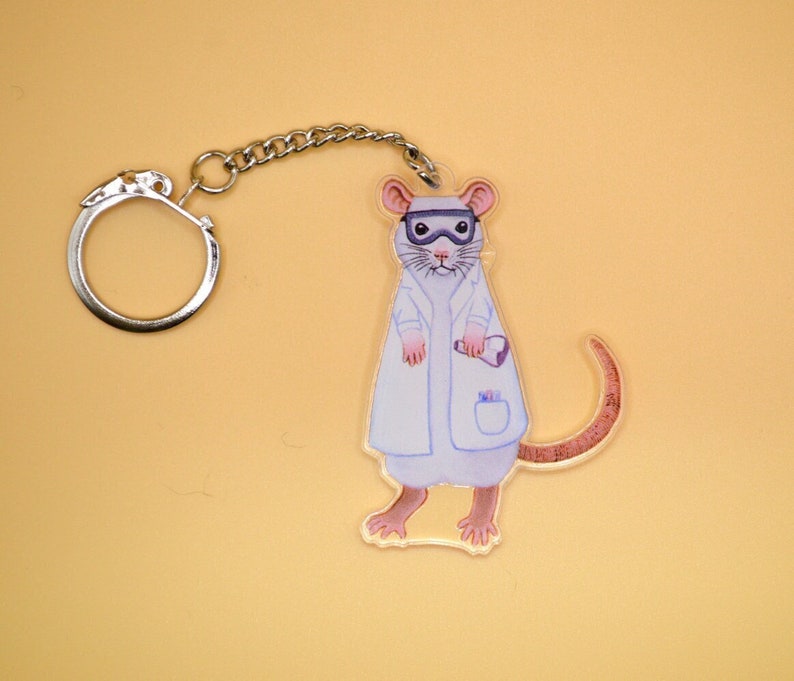 May include: A white lab coat with a pocket and a small beaker is worn by a cartoon rat with a brown tail. The rat is wearing goggles and has a white face. The rat is attached to a silver key chain.