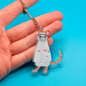 May include: A white lab rat wearing a white lab coat and goggles, holding a test tube. The rat is a keychain made of clear acrylic.