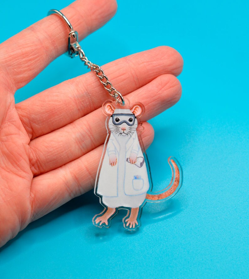 May include: A white lab rat wearing a white lab coat and goggles, holding a test tube. The rat is a keychain made of clear acrylic.