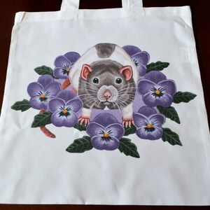 May include: White tote bag with a rat illustration surrounded by purple pansies. The rat has grey and white fur, pink ears and nose. The pansies have purple petals and yellow centres. The bag has white handles.