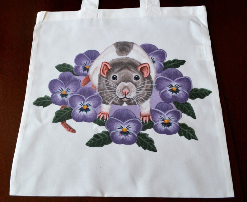 May include: White tote bag with a rat illustration surrounded by purple pansies. The rat has grey and white fur, pink ears and nose. The pansies have purple petals and yellow centres. The bag has white handles.