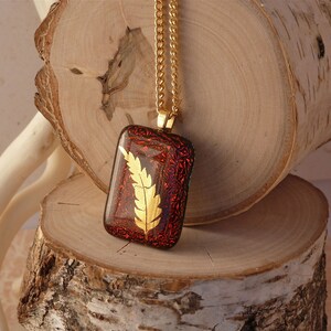 Puede incluir: Un collar de cadena dorada con un colgante de vidrio rojo con un diseño de hoja de oro.