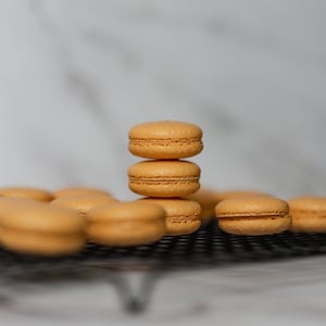 May include: A stack of three orange macarons on a black wire rack. There are other macarons in the background.