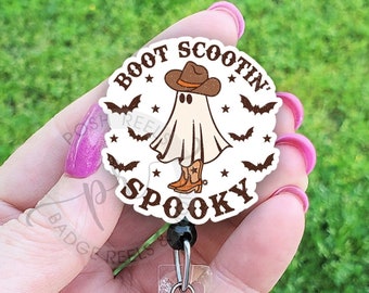 Op de afbeelding: Een wit spook dat een cowboyhoed en laarzen draagt met de tekst "Boot Scootin' Spooky" eromheen. Het spook wordt omringd door vleermuizen en sterren. De afbeelding is op een ronde badge reel.