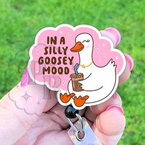Puede incluir: Un ganso de dibujos animados rosa y blanco con una taza de caf&eacute; y el texto "In a silly goosey mood" sobre un fondo rosa en forma de nube. El ganso lleva un collar de oro. La imagen est&aacute; unida a un soporte de credencial retr&aacute;ctil de pl&aacute;stico transparente con un bot&oacute;n negro.