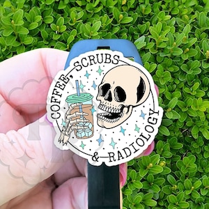 May include: A sticker with the text "COFFEE, SCRUBS & RADIOLOGY" and a skull graphic holding a coffee cup with ice cubes. The sticker is white with black and teal accents, and is placed on a black object.
