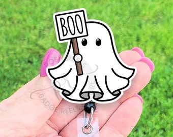 May include: A white ghost badge reel featuring a black outline and a small sign held by the ghost that says BOO in black letters. The badge reel includes a clear plastic strap with a silver metal snap button and a retractable black cord mechanism.