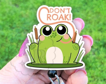 May include: A green frog badge reel with the text "DON'T ROAK!". The frog has large black eyes, a white belly, and is surrounded by green grass and cattails. The badge reel has a clear plastic retractable holder.