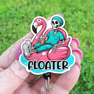 May include: A pink flamingo pool float with a skeleton wearing a blue scrub top and holding a beverage. The float has the word "FLOATER" printed on it. The float is attached to a clear plastic retractable badge holder with a black bead and a silver swivel clip.