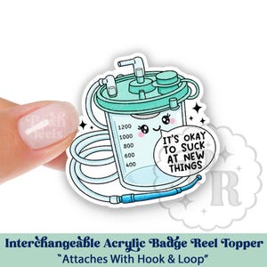 May include: An interchangeable acrylic badge reel topper featuring a cartoon-style medical suction canister with the text "IT'S OKAY TO SUCK AT NEW THINGS." The canister is light blue with a white background.