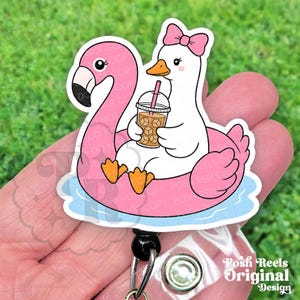 May include: A badge reel featuring a white duck with a pink bow on its head, sitting in a pink flamingo-shaped pool float. The duck holds a clear plastic cup with a pink straw and brown iced beverage. The design has a light blue water base.