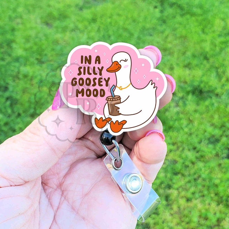 Puede incluir: Un ganso de dibujos animados rosa y blanco con una taza de caf&eacute; y el texto "In a silly goosey mood" sobre un fondo rosa en forma de nube. El ganso lleva un collar de oro. La imagen est&aacute; unida a un soporte de credencial retr&aacute;ctil de pl&aacute;stico transparente con un bot&oacute;n negro.