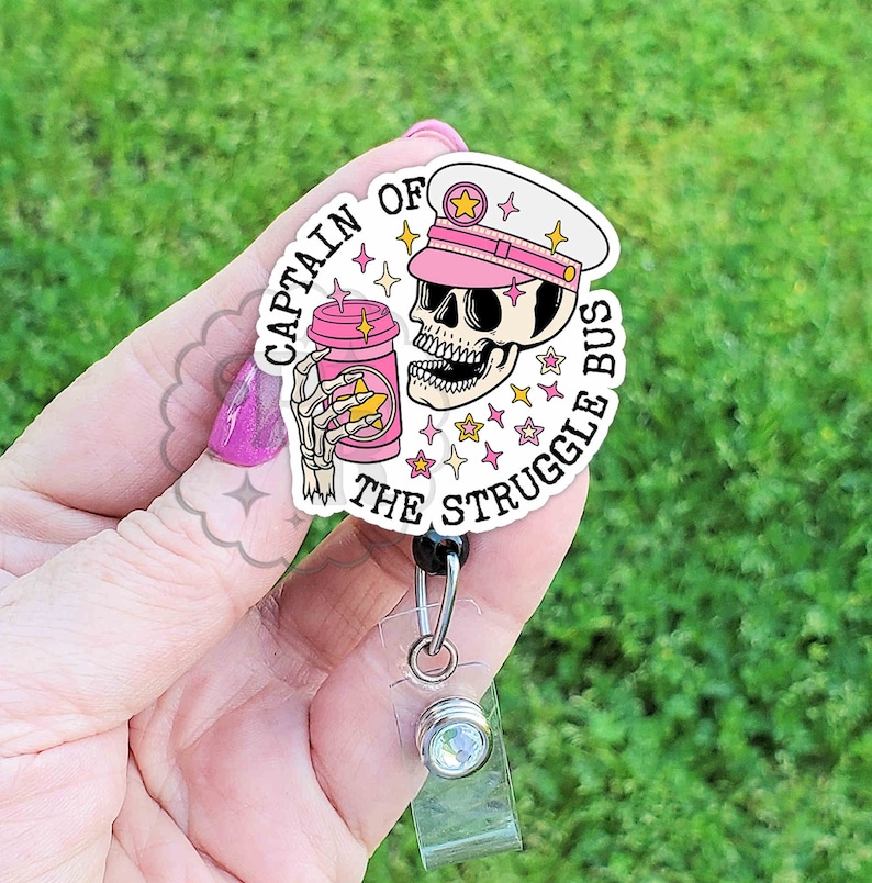 May include: A pink and white badge holder with a cartoon skull wearing a pink captain's hat and holding a pink coffee cup. The text on the badge holder reads "Captain of the Struggle Bus".
