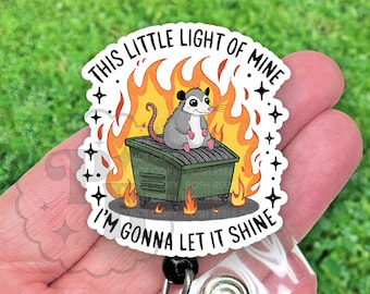 May include: A white and black badge reel with a cartoon opossum sitting in a green trash can on fire. The text on the badge reel reads "This little light of mine I'm gonna let it shine."