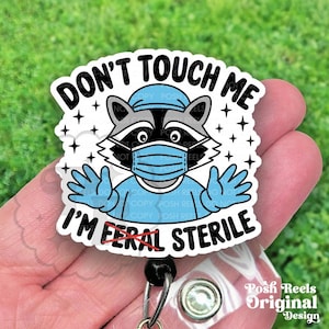 May include: A white badge with a raccoon wearing a blue surgical mask and scrubs. The text "DON'T TOUCH ME" and "I'M FERAL STERILE" are displayed in black. The badge has a retractable reel.