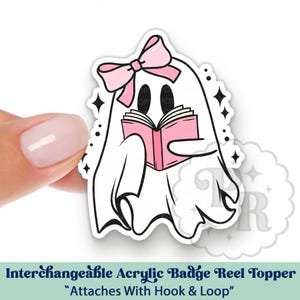 May include: An interchangeable acrylic badge reel topper featuring a white ghost illustration with a pink bow on its head, reading a pink book. The design includes black outlines and small black star accents. The text reads, "Interchangeable Acrylic Badge Reel Topper Attaches With Hook & Loop."