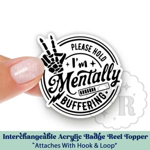 May include: A white and black badge reel topper with the text "PLEASE HOLD I'm Mentally BUFFERING." The design includes a skeleton hand making a peace sign and a loading bar. The topper is held by a person's hand.