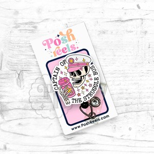 May include: A pink and white badge reel with a skeleton wearing a pink captain's hat and holding a coffee cup. The badge reel has the text "Captain of the Struggle Bus".