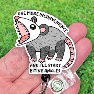 May include: A white and gray opossum badge reel with the text "ONE MORE INCONVENIENCE AND I'LL START BITING ANKLES". The opossum has a pink mouth and tail, and black feet.
