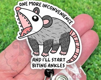 May include: A white and gray opossum badge reel with the text "ONE MORE INCONVENIENCE AND I'LL START BITING ANKLES". The opossum has a pink mouth and tail, and black feet.