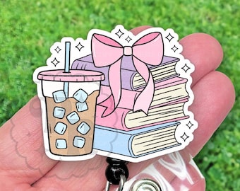 May include: A pink and white cartoon badge reel with a stack of books, a pink bow, and a cup of iced coffee. The badge reel has a clear plastic retractable cord.
