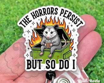 May include: A white and green badge reel with the text "THE HORRORS PERSIST BUT SO DO I". The badge features a cartoon opossum in a dumpster with flames. The opossum is holding a coffee cup.