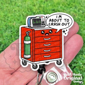 May include: A red medical cart badge reel with a cartoon design. The cart has a monitor, oxygen tank, and the text "I'm about to crash out" in a speech bubble. The badge reel has a clear plastic clip and a metal retractable cord.