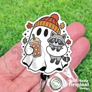 May include: A badge reel featuring a white ghost wearing an orange beanie, holding an iced coffee, and carrying a gray raccoon. The design includes small autumn leaves and sparkles. The text Posh Reels Original Design is visible on the bottom right.