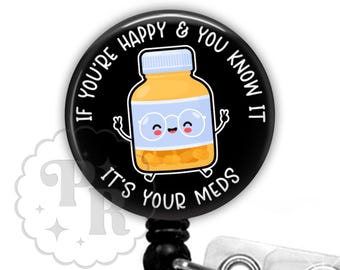 May include: A black badge reel with the text "IF YOU'RE HAPPY & YOU KNOW IT IT'S YOUR MEDS" encircling a cartoon pill bottle wearing glasses. The reel has a black retractable cord and a clear plastic clip with a rhinestone.