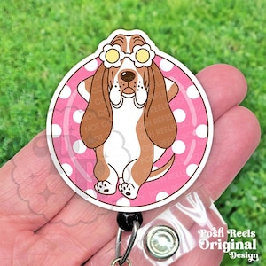 May include: A round, pink and white polka-dotted badge reel featuring a cartoon basset hound wearing flower-shaped sunglasses. The dog is brown, white, and tan. The reel has a black retractable cord and a clear plastic clip. Text on the bottom right reads "Posh Reels Original Design."