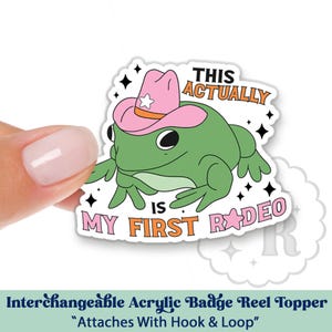 May include: A green frog wearing a pink cowboy hat with a white star. The frog is sitting on a white background with the text "This Actually Is My First Rodeo" in black and orange letters.