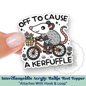 May include: An acrylic badge reel topper with a cartoon opossum on a bicycle, carrying flowers. Text reads "OFF TO CAUSE A KERFUFFLE". The topper attaches with hook and loop. A fun, decorative accessory.