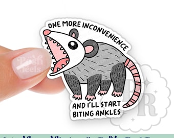 May include: An interchangeable acrylic badge reel topper featuring a cartoon opossum with an open mouth. The opossum is gray with a pink mouth and tail, and the text reads "ONE MORE INCONVENIENCE AND I'LL START BITING ANKLES."