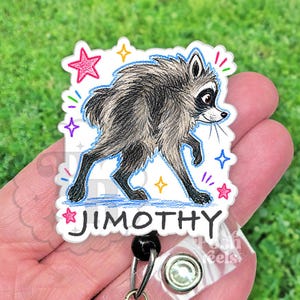 May include: A white acrylic badge reel featuring a colored pencil style illustration of a raccoon walking. The raccoon has grey and black fur. The name JIMOTHY is printed in black capital letters below the raccoon. Colorful stars and marks surround the raccoon.