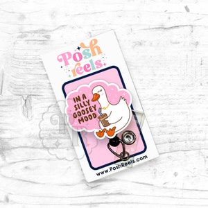 Puede incluir: Una tarjeta rosa y blanca con un ganso de dibujos animados sosteniendo una taza de caf&eacute;. La tarjeta dice "In a silly goosey mood". La tarjeta est&aacute; unida a un portagafete transparente con un carrete retr&aacute;ctil de gafete negro y plateado.