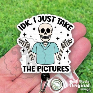 May include: A white badge reel with a cartoon skeleton wearing a teal scrub top. The text "IDK, I JUST TAKE THE PICTURES" is printed in black. The badge reel has a clear plastic base and a black retractable cord.