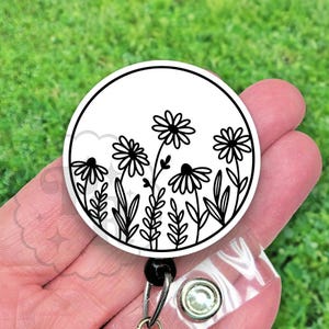 May include: A white round badge holder with a black and white floral design. The design features stylized flowers with five petals and stems with leaves.