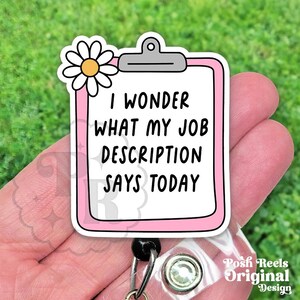 May include: A pink and white badge reel with the text "I WONDER WHAT MY JOB DESCRIPTION SAYS TODAY." The badge reel has a daisy and a silver clip. The words "Posh Reels Original Design" are also visible.