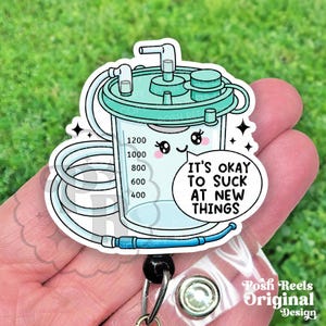 May include: A teal and white badge reel featuring a cartoon illustration of a medical suction canister with the text "IT'S OKAY TO SUCK AT NEW THINGS". The canister has measurement markings and a cute face.