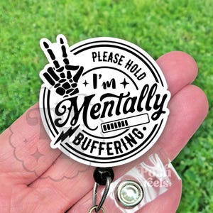 May include: A white and black badge with the text "PLEASE HOLD I'm Mentally Buffering" in a circular design. A black and white skeleton hand makes a peace sign. The badge is held by a clear plastic clip.