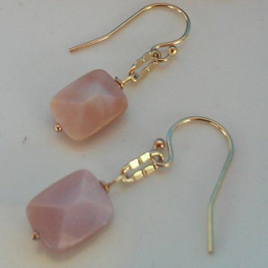 May include: Close-up view of a pair of earrings featuring pale pink, faceted, rectangular stones dangling from gold-toned, delicate chains and hook closures. Three ripe raspberries are in the background, showcasing a color contrast between the jewelry and the fruit. The earrings are displayed on a white surface.