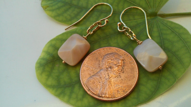 May include: A pair of gold earrings with a light pink square-shaped gemstone. The earrings are hanging from a leaf.