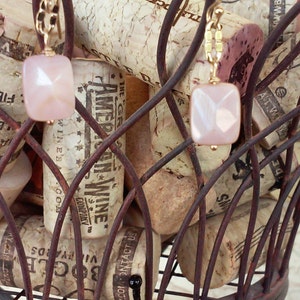 May include: A pair of gold earrings with pink and white gemstone pendants hanging from a wire rack filled with wine corks. The corks have various labels and text, including "The Great American Wine Company" and "Contact Us".