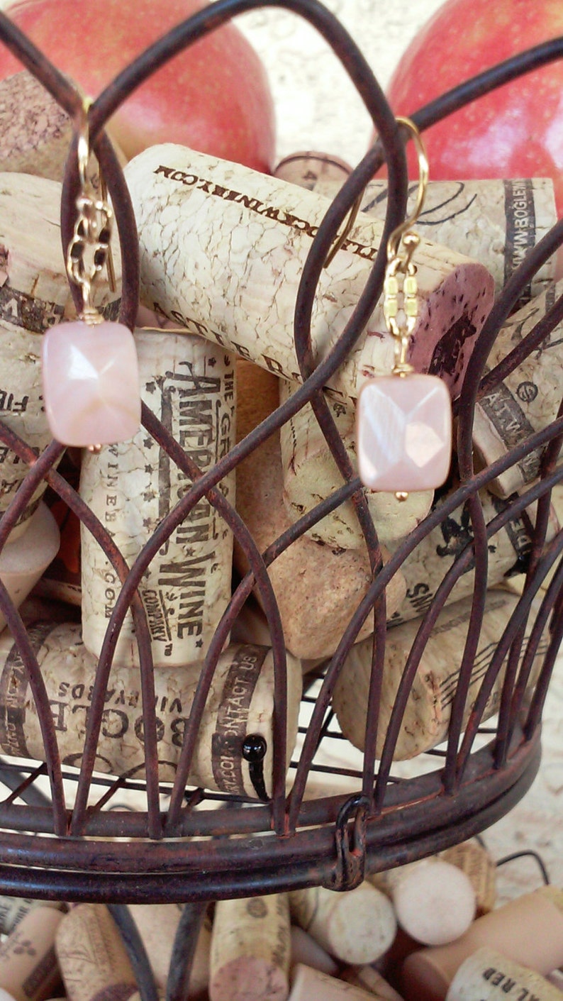 May include: A pair of gold earrings with pink and white gemstone pendants hanging from a wire rack filled with wine corks. The corks have various labels and text, including "The Great American Wine Company" and "Contact Us".