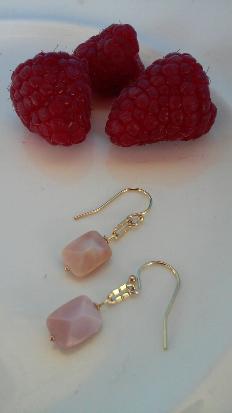 May include: Close-up view of a pair of earrings featuring pale pink, faceted, rectangular stones dangling from gold-toned, delicate chains and hook closures. Three ripe raspberries are in the background, showcasing a color contrast between the jewelry and the fruit. The earrings are displayed on a white surface.