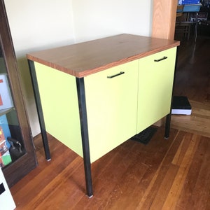 May include: A yellow cabinet with two doors and black metal legs. The cabinet has a wood top and black metal handles.