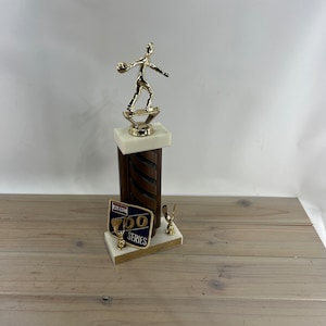 May include: A gold-colored bowling trophy with a wooden base. The trophy features a figure of a bowler throwing a ball. The base has a blue and gold plaque with the text "ABC 700 Series".