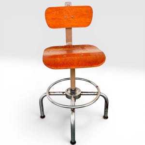 May include: A swivel stool featuring a rectangular wooden backrest and a square wooden seat with rounded corners. The seat and back are supported by a central beige metal post and a circular footrest, resting on four curved metal legs.
