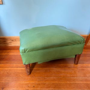 May include: A green leather ottoman with a square top and four wooden legs. The ottoman is a light green color and has a classic design. The legs are a dark brown color and are attached to the base with small metal tacks.