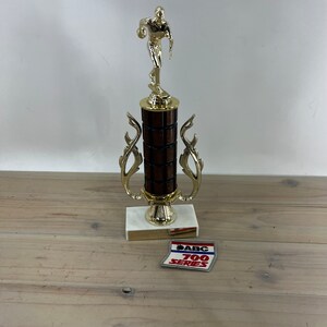 May include: A gold bowling trophy with a brown wooden base and a gold bowler figurine on top. The base has a white marble platform and a gold plaque with the text "ABC 700 SERIES".